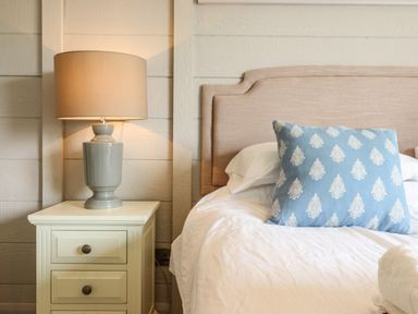 A bedside table with a lamp next to a bed with a beige headboard and a blue decorative pillow at Skylarks in Fornham St Genevieve near Bury St Edmunds
