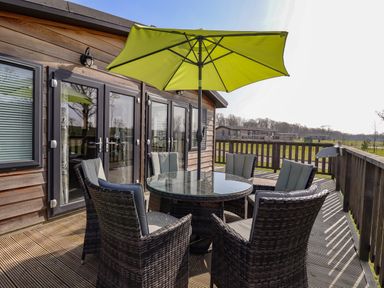 An outdoor wooden deck with a round glass table surrounded by six wicker chairs with cushions and a green patio umbrella at Skylarks in Fornham St Genevieve near Bury St Edmunds