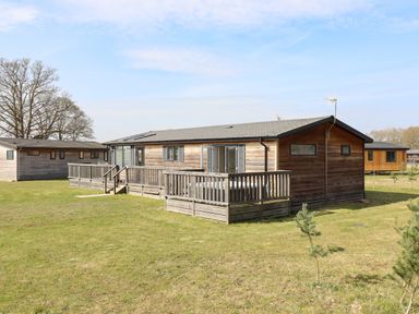 A wooden cabin with a deck and glass doors on a grassy lawn at Skylarks in Fornham St Genevieve near Bury St Edmunds