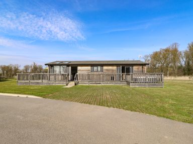 A wooden cabin with a fenced porch situated on grass near a paved road at Skylarks in Fornham St Genevieve near Bury St Edmunds