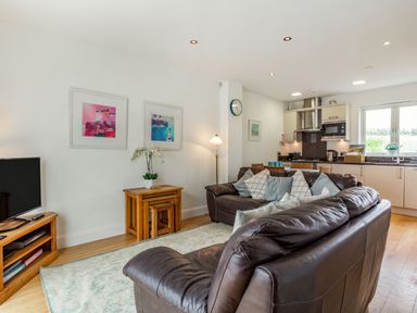 A living room with brown leather sofas a wooden TV stand a side table with nested stools and a kitchen area at Constantine Bay Cottage in Constantine Bay