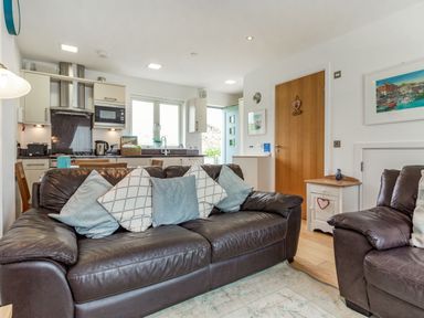An open living room and kitchen with a brown leather sofa and armchair with cushions and kitchen appliances at Constantine Bay Cottage in Constantine Bay