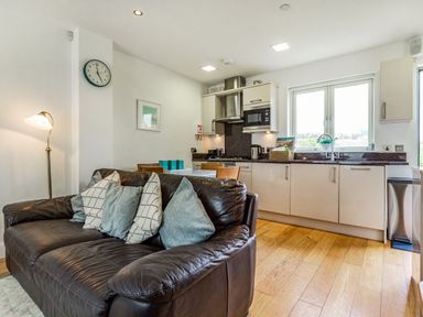 An open plan living area with a dark leather sofa and kitchen with white cabinets and wooden floor at Constantine Bay Cottage in Constantine Bay