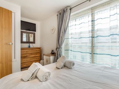 A bedroom with a bed covered with white sheets and rolled towels a wooden dresser a wall-mounted television and a window with sheer striped curtains at Constantine Bay Cottage in Constantine Bay