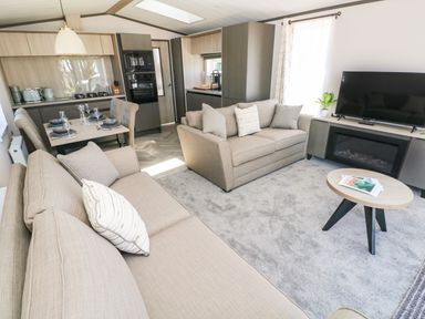 A living room with two beige sofas a flat screen tv on a cabinet a small round coffee table and a dining table and kitchen area at Puffin Lodge in Broad Haven