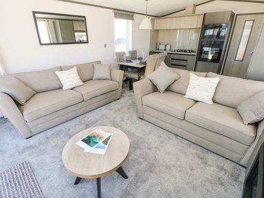 A living room with two beige sofas a round wooden coffee table a dining area and a kitchen in the background at Puffin Lodge in Broad Haven