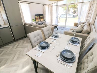 A dining table set for four with plates and glasses in a living room with sofas and a television and sliding glass doors at Puffin Lodge in Broad Haven