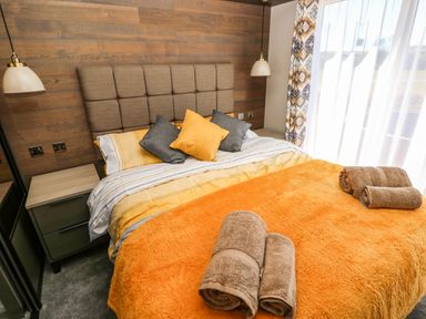 A bedroom with a double bed with orange and gray pillows and blankets beside a nightstand and window at Puffin Lodge in Broad Haven