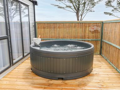 A round hot tub on a wooden deck with a bamboo fence and a bottle of champagne with glasses at Puffin Lodge in Broad Haven