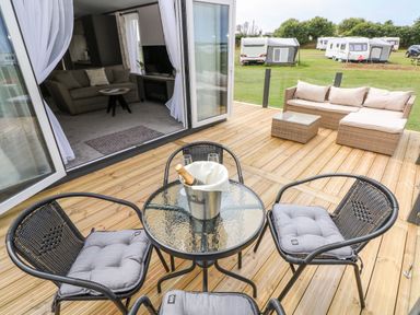 A patio with a round glass table and four chairs with cushions next to an open living room with a sofa and a coffee table at Puffin Lodge in Broad Haven