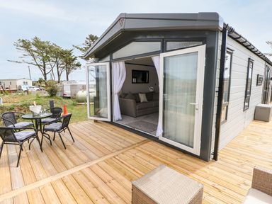 An outdoor wooden deck with a glass table and four chairs next to a small building with open double doors at Puffin Lodge in Broad Haven