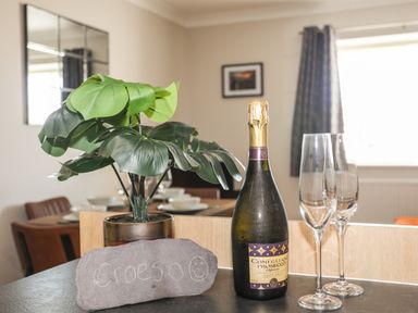 A kitchen counter with a potted plant a bottle of prosecco two empty champagne glasses and a stone with writing in a dining area at 4 Pen Llanw Tides Reach in Rhosneigr