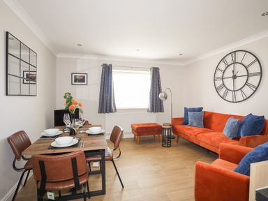 A living and dining area with a wooden dining table set with dishes and glasses next to an orange sofa with blue cushions and a large wall clock at 4 Pen Llanw Tides Reach in Rhosneigr