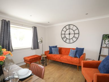 A living room with orange sofas and blue cushions a large wall clock and a window with curtains at 4 Pen Llanw Tides Reach in Rhosneigr