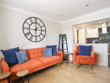A living room with orange sofa and armchair with blue cushions and large black clock on wall at 4 Pen Llanw Tides Reach in Rhosneigr