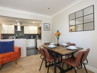 A dining area with a wooden table set with plates and glasses next to an open kitchen with light cabinets and black backsplash at 4 Pen Llanw Tides Reach in Rhosneigr