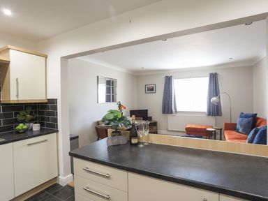 A kitchen counter with a bottle and two glasses looking into a living room with orange sofa and blue pillows at 4 Pen Llanw Tides Reach in Rhosneigr