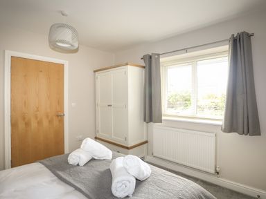 A bedroom with a wooden door a white wardrobe gray curtains and rolled white towels on a gray bedspread at 4 Pen Llanw Tides Reach in Rhosneigr