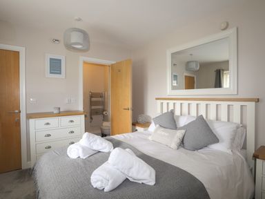 A bedroom with a bed with gray and white bedding and rolled towels with a dresser and an open door showing a bathroom at 4 Pen Llanw Tides Reach in Rhosneigr