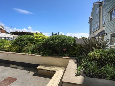 An outdoor garden area with plants bushes a wooden bench and residential buildings in the background at 4 Pen Llanw Tides Reach in Rhosneigr