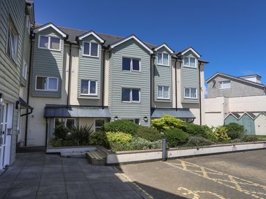 An exterior view of a residential building with multiple windows shrubs in front and a paved parking area at 4 Pen Llanw Tides Reach in Rhosneigr