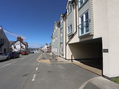 A street with parked cars and buildings including a covered parking area at 4 Pen Llanw Tides Reach in Rhosneigr