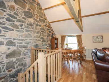A dining area with wooden chairs and table next to a window with curtains a stone wall a wooden staircase railing and a brown leather sofa at Bryn Moelyn Cottage in Yr Efail near Morfa Nefyn