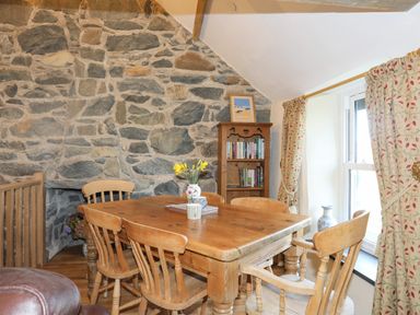 A dining area with a wooden table and chairs beside a stone wall and window with patterned curtains at Bryn Moelyn Cottage in Yr Efail near Morfa Nefyn