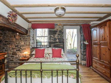 A bedroom with a metal bed frame red pillows floral bedding wooden wardrobe wooden floor and exposed stone walls at Bryn Moelyn Cottage in Yr Efail near Morfa Nefyn