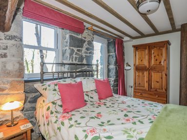 A bedroom with a floral bedspread and pink pillows a wooden wardrobe and stone walls at Bryn Moelyn Cottage in Yr Efail near Morfa Nefyn