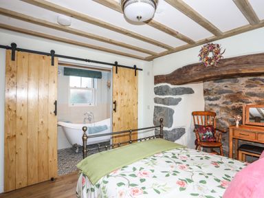 A bedroom with a floral bedspread a wooden chair a dresser and sliding wooden doors opening to a bathroom with a bathtub at Bryn Moelyn Cottage in Yr Efail near Morfa Nefyn