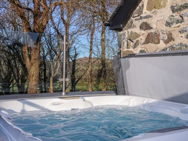 A hot tub with bubbling water next to a stone wall and glass railing with trees in the background at Bryn Moelyn Cottage Yr Efail near Morfa Nefyn