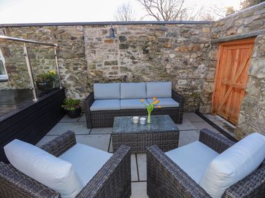 An outdoor patio with wicker sofa and chairs around a glass table with flowers and mugs against a stone wall at Bryn Moelyn Cottage in Yr Efail near Morfa Nefyn
