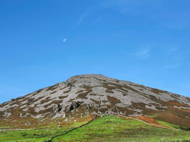 A hill with scattered rocks and patches of green grass under a blue sky with the moon visible