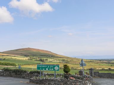 A green sign for Tafarn Y Fic near a stone wall and rural landscape with hills in the background at Bryn Moelyn Cottage in Yr Efail near Morfa Nefyn