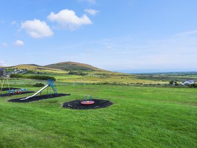 A playground with a slide swings and a roundabout on a grassy field with hills in the background at Bryn Moelyn Cottage in Yr Efail near Morfa Nefyn