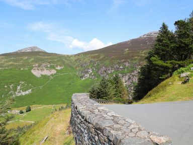 A winding mountain road with a stone wall and trees beside green hills and peaks at Bryn Moelyn Cottage in Yr Efail near Morfa Nefyn