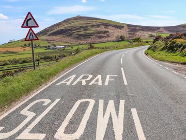 A curved road with a slow and araf marking and road signs showing a right bend and road narrowing with hills and fields in the background at Bryn Moelyn Cottage in Yr Efail near Morfa Nefyn