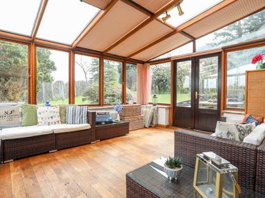 A sunroom with wicker sofas with cushions a glass coffee table and large windows overlooking a garden at Gilfachwydd Islawrdref near Dolgellau