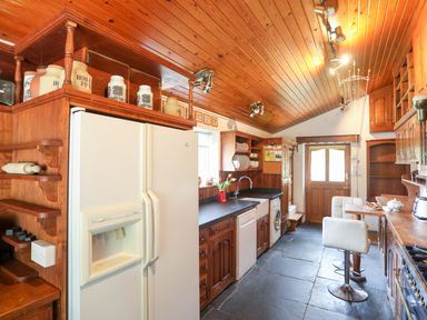A kitchen with wooden cabinets a white refrigerator dishwasher and washing machine with a countertop and bar stools at Gilfachwydd in Islawrdref near Dolgellau
