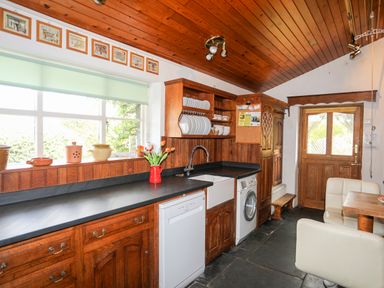 A kitchen with wooden cabinets a black countertop white dishwasher and washing machine a wooden door and chairs at Gilfachwydd Islawrdref near Dolgellau