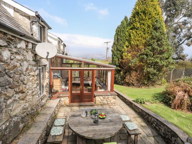 An outdoor patio area with a round wooden table and benches with cushions next to a stone house with a glass conservatory and garden at Gilfachwydd Islawrdref near Dolgellau
