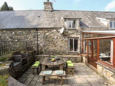 An outdoor patio area with a round wooden table and benches with cushions next to a stone house with a glass conservatory at Gilfachwydd in Islawrdref near Dolgellau