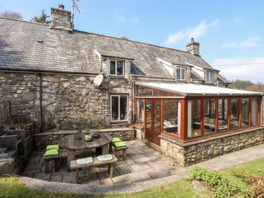 An outdoor patio with a round wooden table and benches next to a stone house with a glass sunroom at Gilfachwydd Islawrdref near Dolgellau