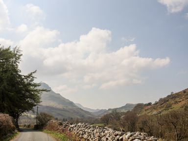 A rural road with stone walls and trees leading towards mountains under a partly cloudy sky at Gilfachwydd in Islawrdref near Dolgellau