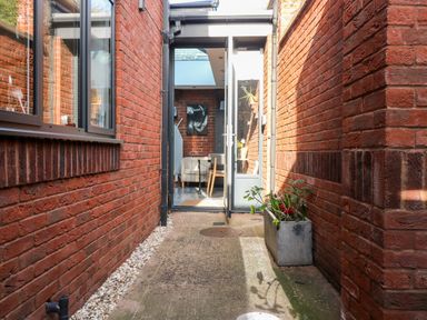 A narrow outdoor corridor with red brick walls on both sides a concrete planter with plants and an open glass door leading to a room with chairs at Farriers Retreat Budleigh Salterton