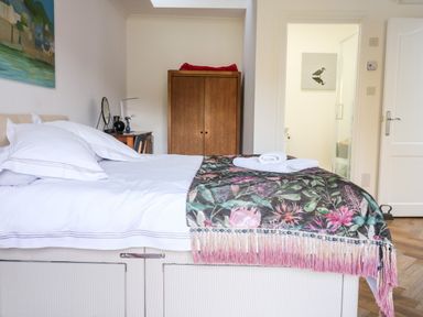 A bedroom with a bed covered with white sheets and a floral patterned blanket with towels on top a wooden wardrobe a desk with a chair and a mirror and an open door leading to a bathroom at Farriers Retreat in Budleigh Salterton
