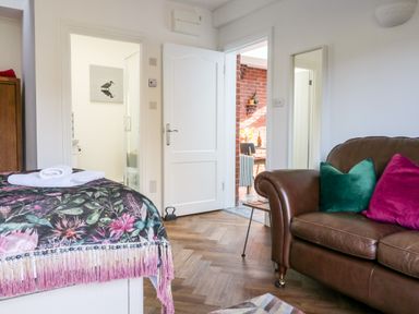 A bedroom with a bed with a floral blanket and white towels a brown leather sofa with green and magenta cushions and an open door showing a bathroom and a brick wall in the next room at Farriers Retreat Budleigh Salterton