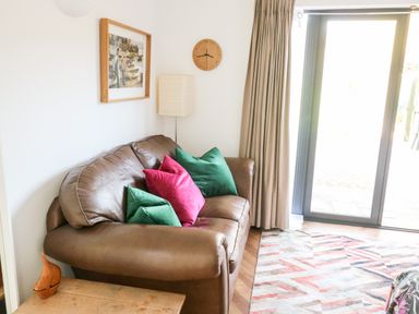 A brown leather sofa with green and red cushions next to a window with beige curtains at Farriers Retreat in Budleigh Salterton