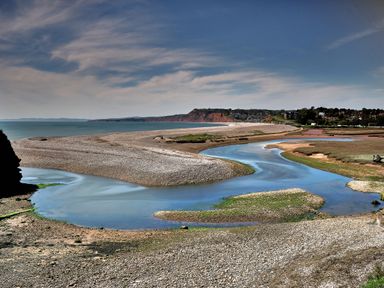 A river winding through a pebble beach landscape with a coastal town and cliffs in the background at Farriers Retreat in Budleigh Salterton